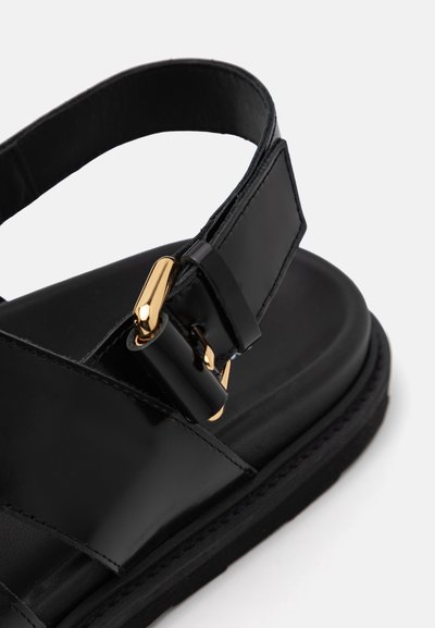 Black sandal with a glossy leather finish, featuring a gold buckle and a wide strap design for added support. Textured sole for grip.