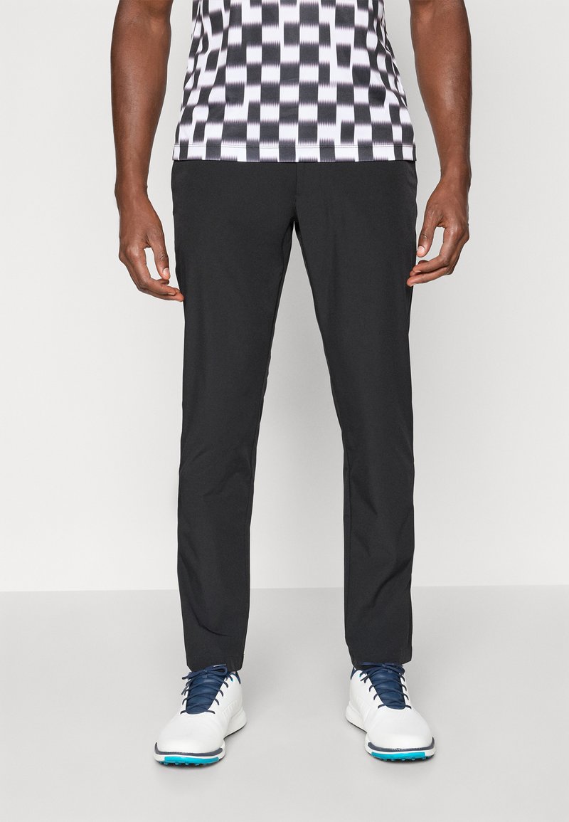 Pantalon noir ajusté en tissu léger ; texture lisse ; coupe slim ; associé à une chemise à motif et des chaussures de sport blanches.