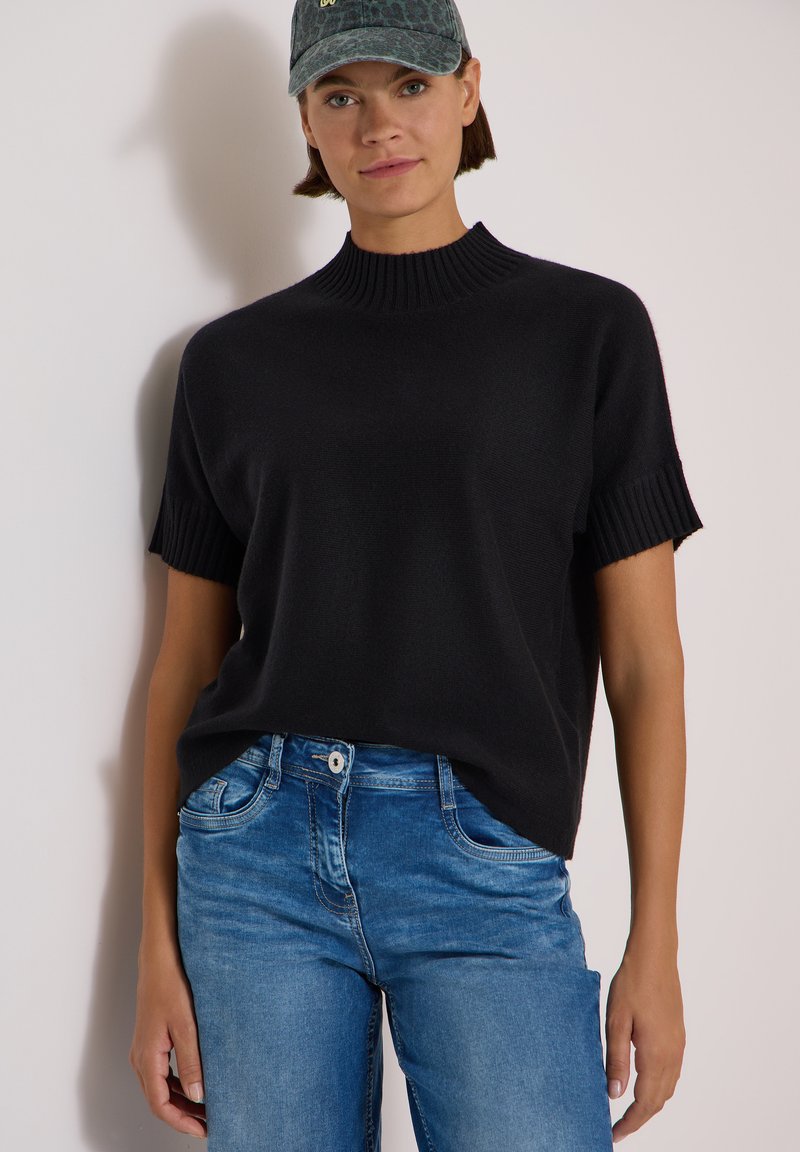 Femme portant un pull noir à manches courtes et col montant, un jean bleu et une casquette vert foncé sur un fond clair uni.