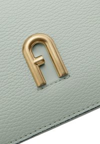 Dettaglio di una borsa in pelle verde chiaro con un logo in metallo dorato prominente che assomiglia alla lettera "f" e cuciture in vista.