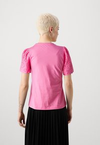 Pink short-sleeve t-shirt featuring eyelet detail on sleeves, round neckline, and a relaxed fit, worn with a pleated black skirt.