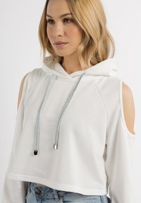 Felpa bianca corta con maniche lunghe e design off-the-shoulder. Presenta un cordoncino con accenti metallici. Texture morbida e liscia.