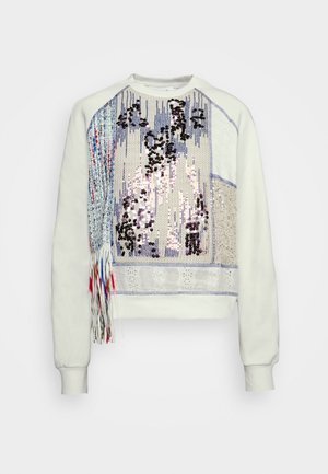 Sweat-shirt blanc avec des patchs à sequins de différentes couleurs et textures, présentant un détail à franges sur un côté.