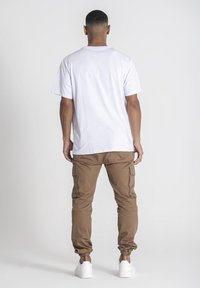 Camiseta blanca de manga corta y pantalones cargo marrones con puños elásticos, que cuentan con bolsillos en los muslos. El modelo lleva zapatillas blancas.