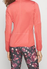 Veste de sport corail avec des détails texturés et des manches longues, associée à des leggings noirs ornés d'un motif floral rose et bordeaux.