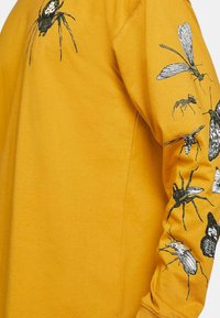 Hanorac galben cu ilustrații detaliate de insecte negre pe mâneci. Materialul pare moale, cu o croială lejeră și decolteu rotund.