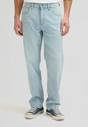 Jeans azzurri diritto indossati da una persona in piedi, abbinati a sneakers blu navy e bianche su uno sfondo semplice.