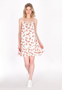 Abito bianco con motivo a ciliegie rosse e verdi, orlo svasato, spalline regolabili e tessuto leggero. Indossato con sneaker bianche.