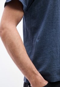 Δεξί χέρι ενός ατόμου που φοράει μπλε κοντομάνικο πουκάμισο, με το χέρι του να ακουμπά στην τσέπη σκούρου παντελονιού, πάνω σε απλό φόντο.