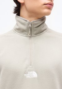 Νεαρό άτομο που φοράει ανοιχτό γκρι φλις μπλούζα με μισό φερμουάρ και το λογότυπο The North Face στο στήθος.