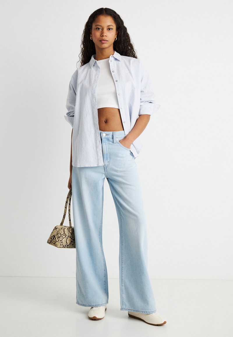 Jeune femme aux cheveux bouclés portant un jean large bleu clair, un crop top blanc, une chemise rayée ouverte, des chaussures blanches et tenant un sac à main imprimé serpent.