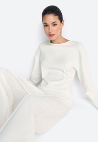 Valge pika varrukaga pluus ümarate kaeluse, pehme tekstuuri ja minimaalsete õmblustega, kombineeritud laia lahuga pükstega, kõik ühtlases heledas toonis.