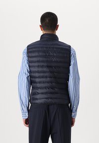 Marineblauwe puffer vest met verticale gestikte ontwerp, hoge kraag en ritssluiting, gedragen over een lichtblauw gestreept overhemd.
