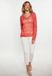 Koralowy sweter dzianinowy z dekoltem w serek, z zygzakowatym wzorem i otwartym splotem. W zestawie z białymi slim-fit spodniami i jasnymi zamszowymi loafersami.