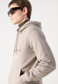 Felpa beige con tasca frontale, caratterizzata da un grande logo bianco a "K" e testo dorato. Occhiali da sole rettangolari neri, modello con capelli corti.