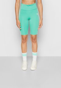 Person som bär turkosa Adidas högmidjade cykelbyxor, vita strumpor med turkosa ränder och vita Adidas sneakers, som står mot en vit vägg.