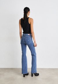 Woman with long dark hair in a ponytail wears a black sleeveless top, high-waisted blue jeans, and black heeled boots, standing against white wall.