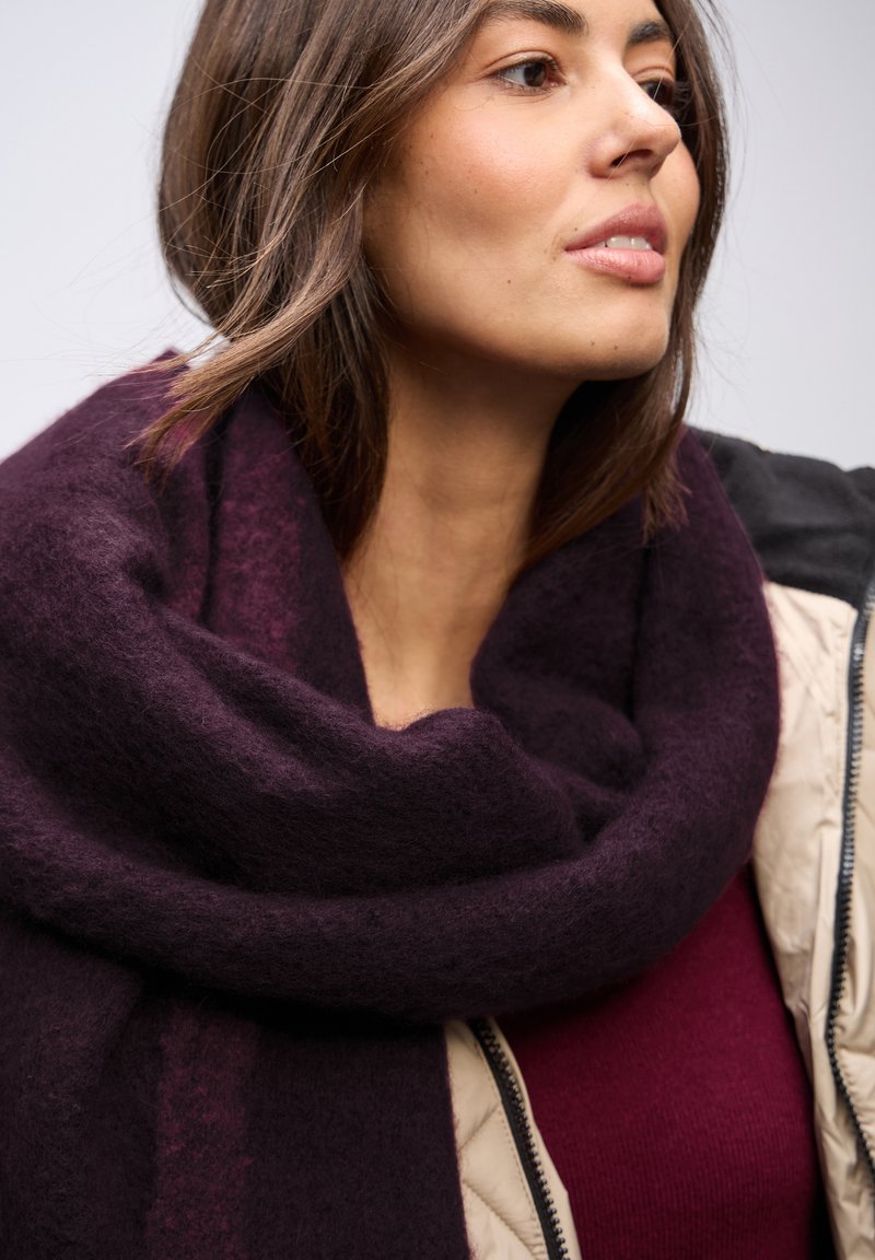 Écharpe en velours de couleur violet foncé au toucher doux, drapée élégamment autour du cou, assortie à un haut bordeaux profond et une veste de couleur claire.