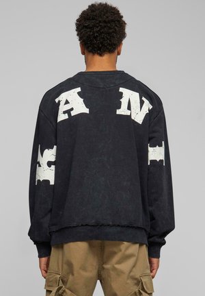 Sudadera negra de gran tamaño con letras blancas en un acabado desgastado en la parte trasera y las mangas. Presenta puños y dobladillo acanalados, y una textura desgastada.