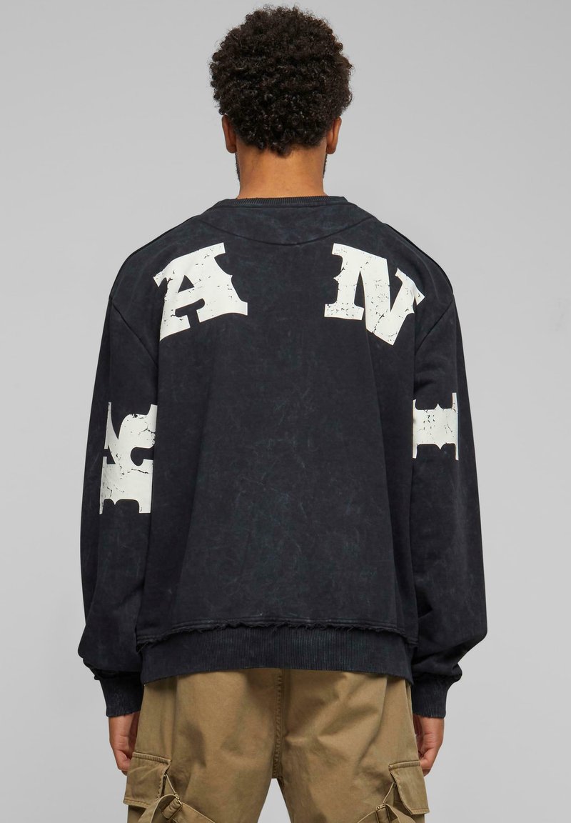 Zwarte oversized sweatshirt met witte versleten letters op de achterkant en mouwen. Heeft ribgebreide manchetten enzoom, en een versleten textuur.