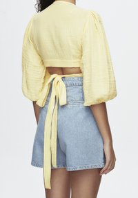 Haut court jaune à manches bouffantes avec un détail noué dans le dos, associé à un short en denim bleu clair. Le haut est en tissu texturé.