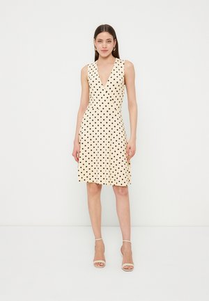 Femeie în picioare, purtând o rochie crem fără mâneci cu buline negre și decolteu în V, sandale beige cu toc înalt și cercei aurii de tip cerc.