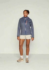 Γυναίκα που στέκεται φορώντας μια γκρίζα ζακέτα με κουκούλα, μπεζ σορτς, λευκές κάλτσες και μπεζ αθλητικά παπούτσια σε ένα απλό ανοιχτό φόντο.