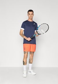 Granatowa koszulka sportowa z białymi rękawami, w połączeniu z żywymi pomarańczowymi szortami. Trzyma rakietę tenisową, widoczne białe sneakersy oraz skarpetki.