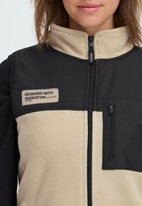 Schwarwest und beiger Reißverschluss-Westen mit einer strukturierten Fleece-Front und einem glatten Rückenteil. Verfügt über eine kleine Reißverschlusstasche und ein Etikett mit der Aufschrift "Designed with Dedication."