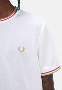Camisa polo blanca con textura de malla, que presenta un logotipo bordado en oro y acentos de rayas de colores en el cuello y las mangas.