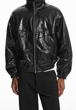 Schwarze glänzende, strukturierte Zip-Up-Jacke mit Krokodilmuster, getragen mit schwarzer Hose, zeigt Vorderansicht und Oberkörper.