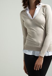 Suéter beige con escote en V y mangas largas, con detalles de herrajes dorados en los hombros, superpuesto a una camisa blanca con cuello.