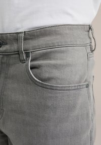 Sive teksas hlače z oprijeto zasnovo, ki vključujejo sprednje žepe, zanke za pas ter črne gumbne detajle. Gladka tekstura, šivani šivi.