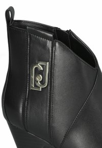 Stivale alla caviglia in pelle nera con finitura lucida, dotato di zip laterale e un accento con logo argento. Il design presenta linee eleganti e curvate.