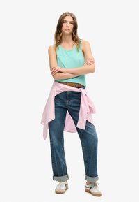 Top in verde menta, camicia rosa chiaro annodata in vita, jeans in denim blu larghi e sneaker bianche con dettagli neri. Outfit casual.