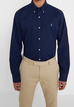 Mand iført en mørkeblå skjorte med knapper og lange ærmer med lyst blåt logo og beige bukser mod en ensfarvet baggrund.