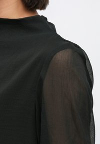 Μαύρο τοπ με ψηλό λαιμό και διάφανο μαύρο ύφασμα στους ώμους, με απαλή υφή και ελαφριά γυαλάδα.