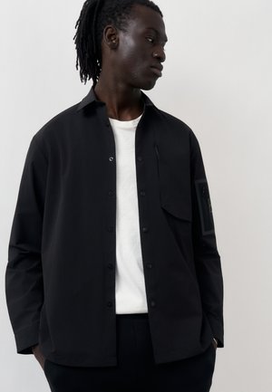 Mężczyzna o ciemnej karnacji z dredami nosi czarną kurtkę z guzikami na białej koszuli, stoi z rękami w kieszeniach, patrząc w prawo.