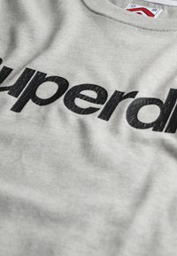 Grå sweatshirt lavet af en bomuldsblandingsstof med et stort, blankt sort "superdry" logo tværs over brystet i en tekstureret finish.