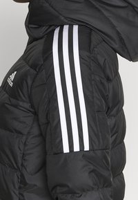 Černá péřová bunda s bílými pruhy Adidas na ramenou. Materiál vypadá leskle a má prošívanou texturu.