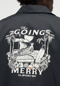 Spatele unei jachete negre cu design tropical alb, palmieri, navă, placă de surf, pălărie și textul „The Going Merry” și „Quiksilver.”
