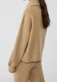 Beżowy sweter dziewiarski z wysokim kołnierzem, luźny krój i prążkowane mankiety, w połączeniu z pasującymi, teksturowanymi spodniami. Miękka, lekka tkanina.