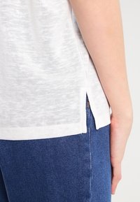 Top blanco de manga corta con una textura ligeramente transparente, con aberturas en los laterales. Combinado con jeans de mezclilla azules.
