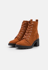 Bottines en daim marron avec bout arrondi, design à lacets, œillets argentés et talon noir épais. Semelle en caoutchouc texturée pour une meilleure adhérence.