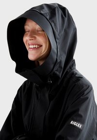 Veste imperméable noire avec une grande capuche, dotée d'une fermeture éclair à l'avant et d'une patch logo "AIGLE" sur la manche. Texture lisse et mate.