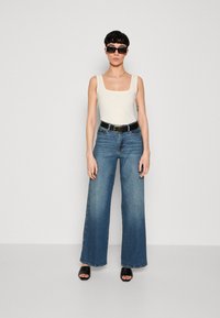 Jean en denim bleu à jambes larges avec un débardeur ajusté beige clair, une ceinture noire et des sandales à talons ouverts noires. Des lunettes de soleil carrées complètent le look.