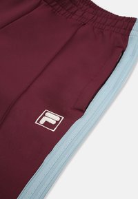 Mörkröda sportbyxor med ljusblå sidokanter. Har en elastisk midja och en vit broderad logotyp på vänster ben.