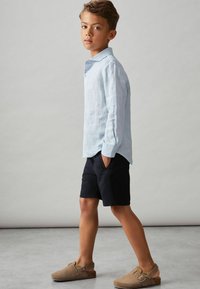 Ljusblå linneskjorta, svarta shorts och bruna slip-on skor. Skjortan har krage och knappdetaljer, med en avslappnad passform.