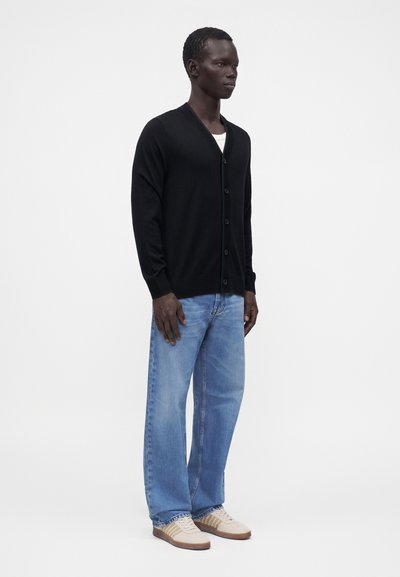 Homme debout portant un cardigan noir boutonné, un jean bleu clair et des baskets beige sur fond blanc uni.