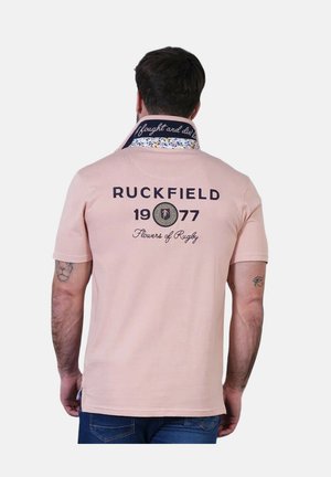 Homme portant un polo rose clair avec le texte « RUCKFIELD 1977 Flowers of Rugby » au dos, tourné de dos montrant des tatouages sur les bras.